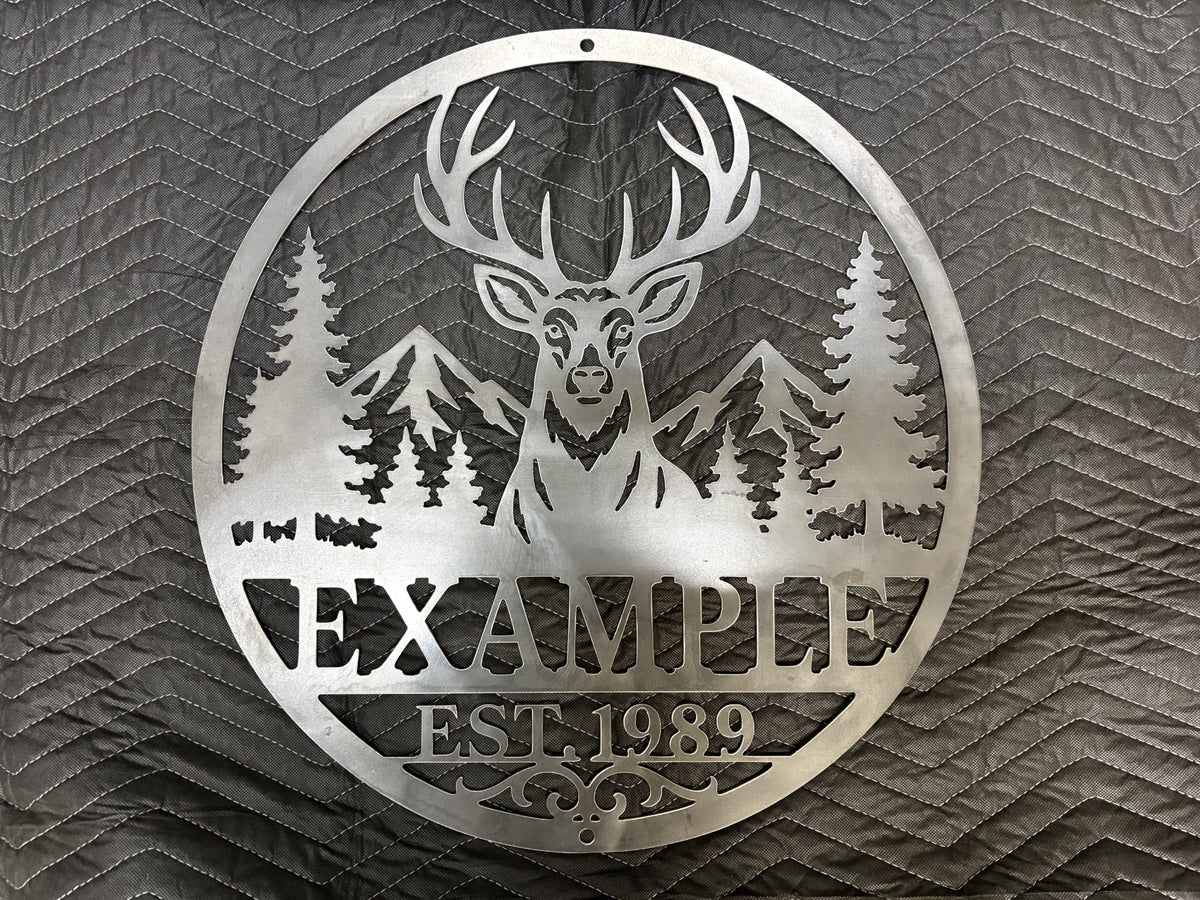 A circular metal wall sign featuring a deer, mountains, pine trees, and the text 'EXAMPLE EST. 1989', shown against a dark textured background.